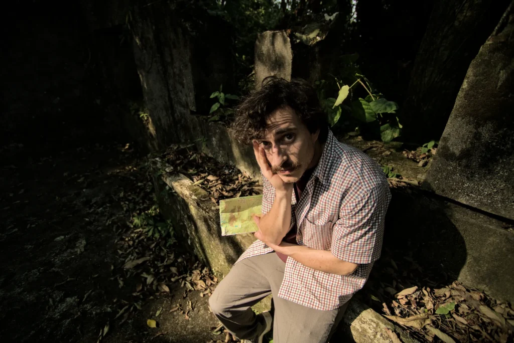 A man looks at the camera while sitting in the ruins of an old home in the jungle. He is lost and desperate.